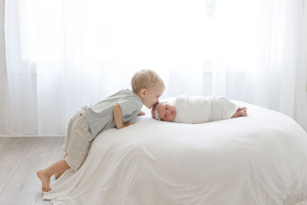 A young boy kisses the forehead of his newborn baby sister lying in a white swaddle in a studio liz lehmann