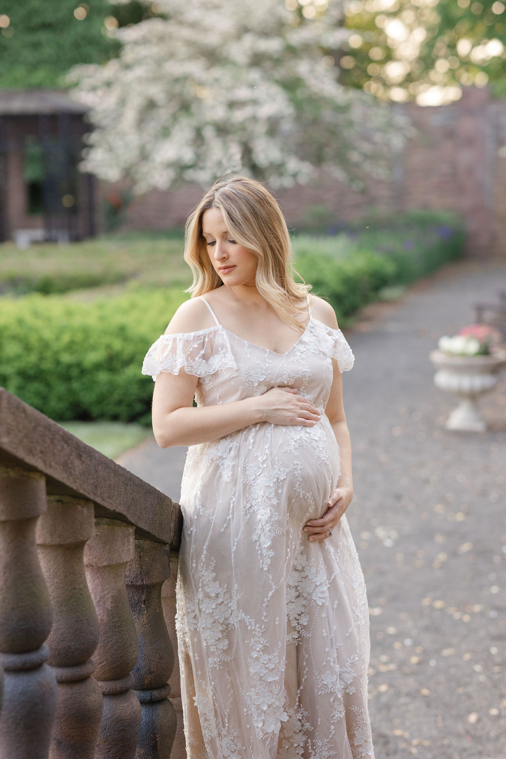 A mom to be stands at the bottom of some stairs in a garden while looking down her shoulder and holding her bump
