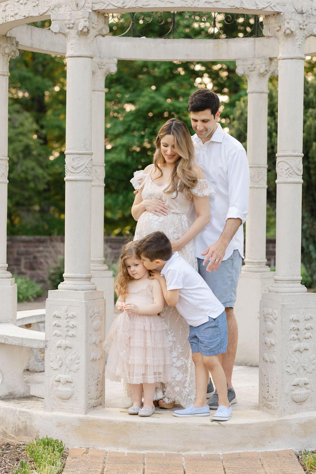 A pregnant mother leans into her husband while their young son kisses his little sister on the cheek while standing under a garden arch prenatal fitness philadelphia