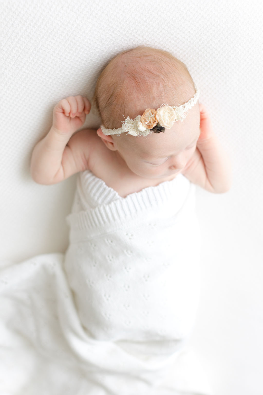 A newborn baby sleeps in a white blanket wearing a floral headband