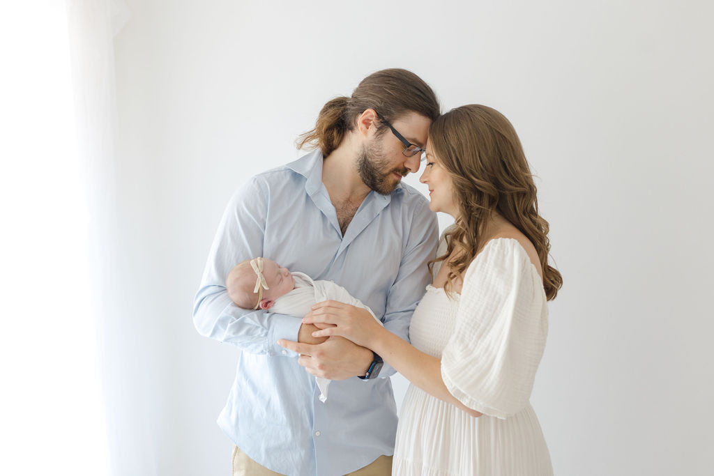 New parents touch foreheads while standing in a studio holding their sleeping newborn baby baby supermart broomall pa