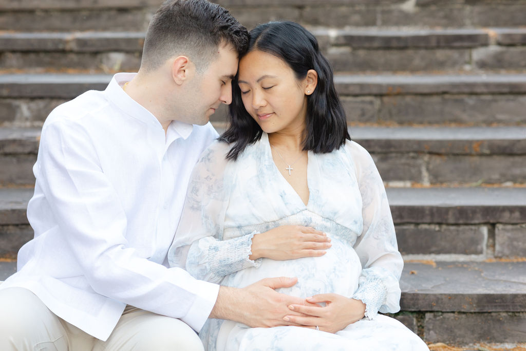 Expecting parents touch foreheads while placing hands on the bump and sitting on stone stairs
