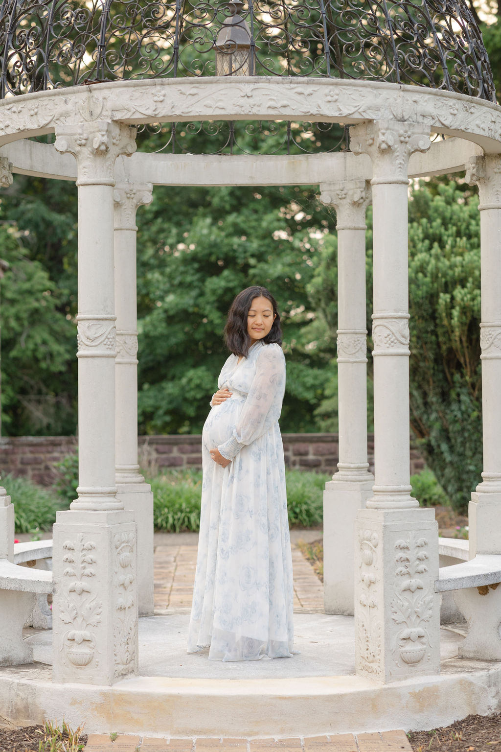 A mom to be in a blue maternity dress looks down her shoulder while standing in a garden gazebo philadelphia prenatal massage
