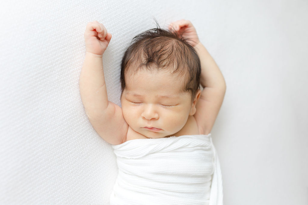 A newborn baby stretches arms above its head while sleeping in a white wrap