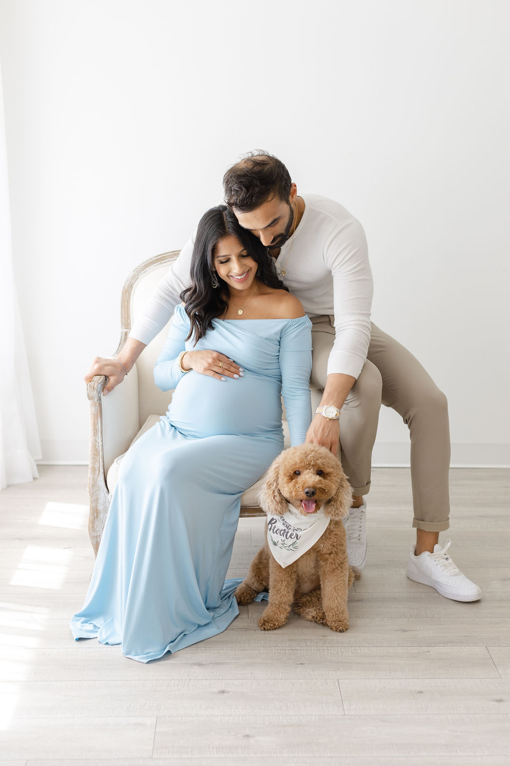 A mom to be sits in a chair with her husband on the arm in a blue maternity gown with their fluffy dog at their feet philadelphia baby shower venues
