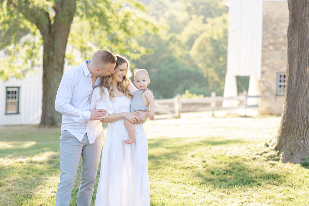 A mother and father stand in a park wearing a white dress and button down shirt holding and playing with their one year old baby on mom's hip at sunset