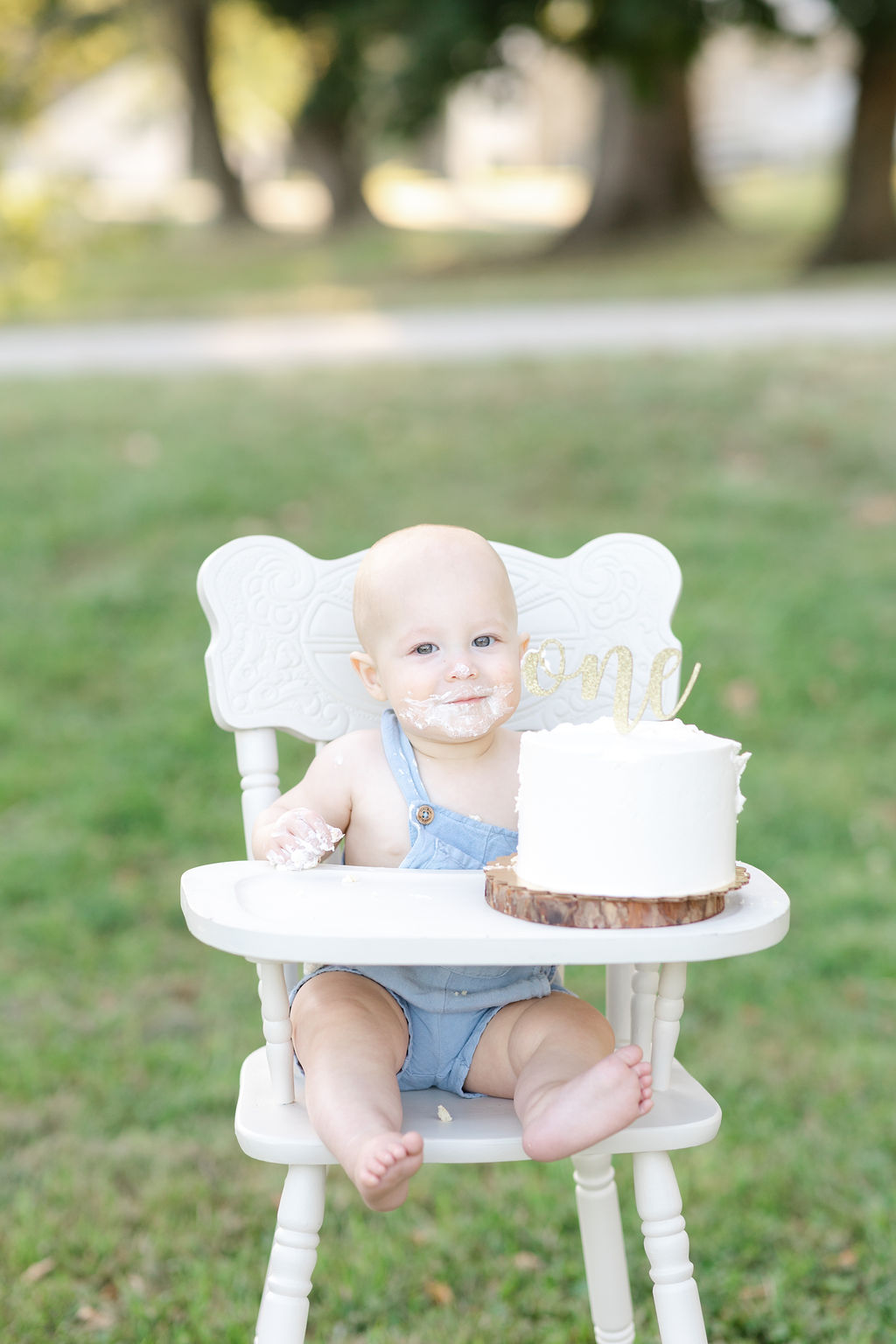 A one year old sits in a white wooden high chair in a park covered in cake from a bakery in Newtown PA