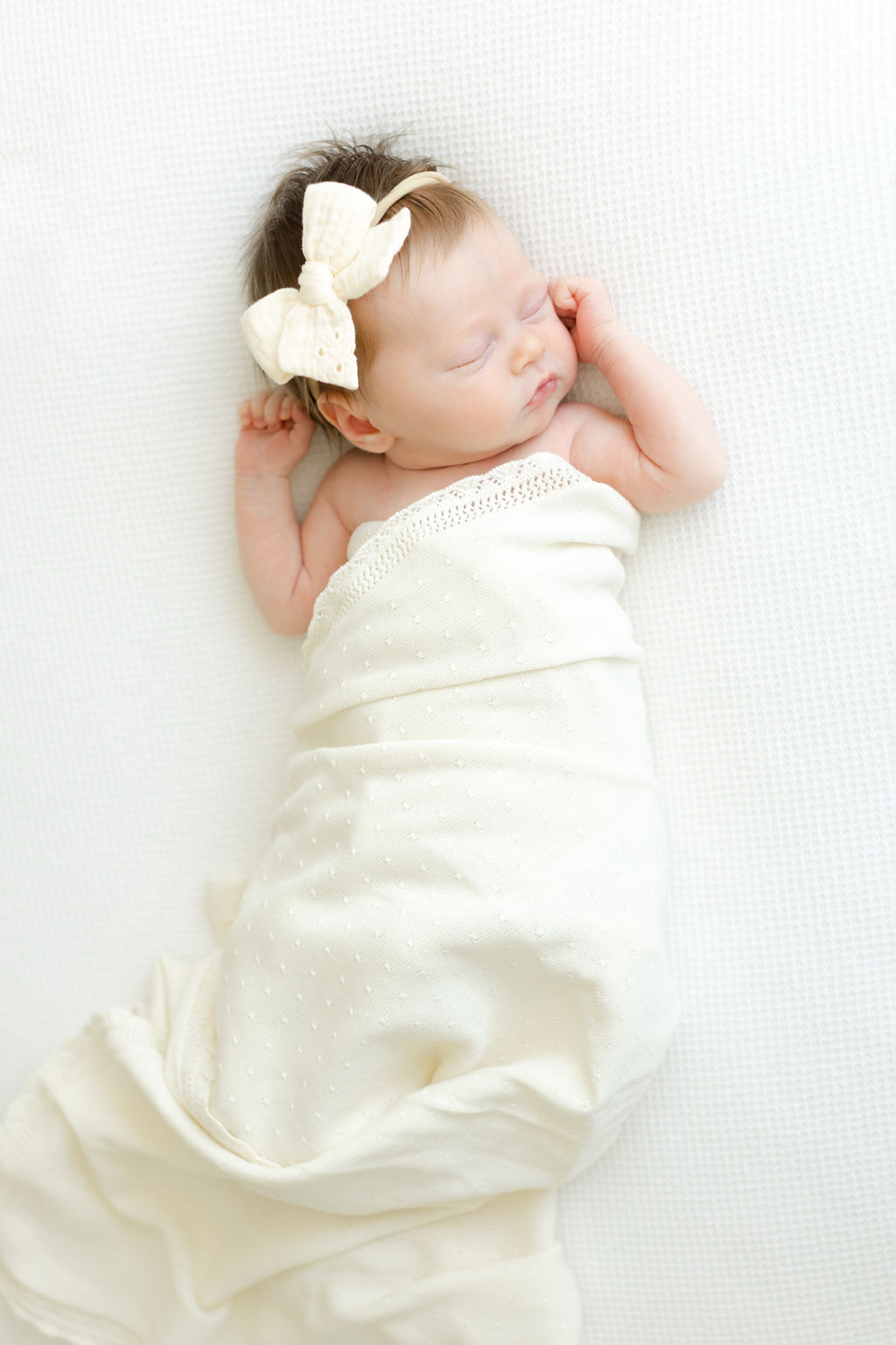 A newborn baby sleeps in a cream blanket and matching bow on a white bed