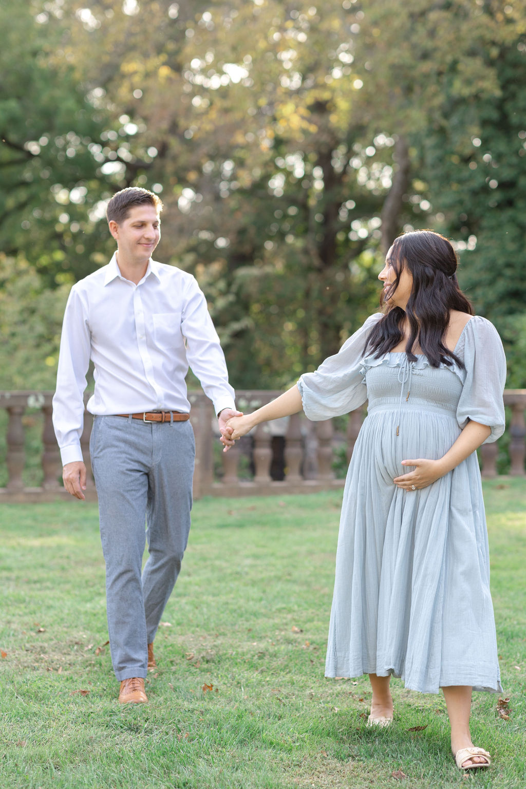 A mom to be in a blue maternity gown leads her partner in a white shirt and grey slacks through a park at sunset