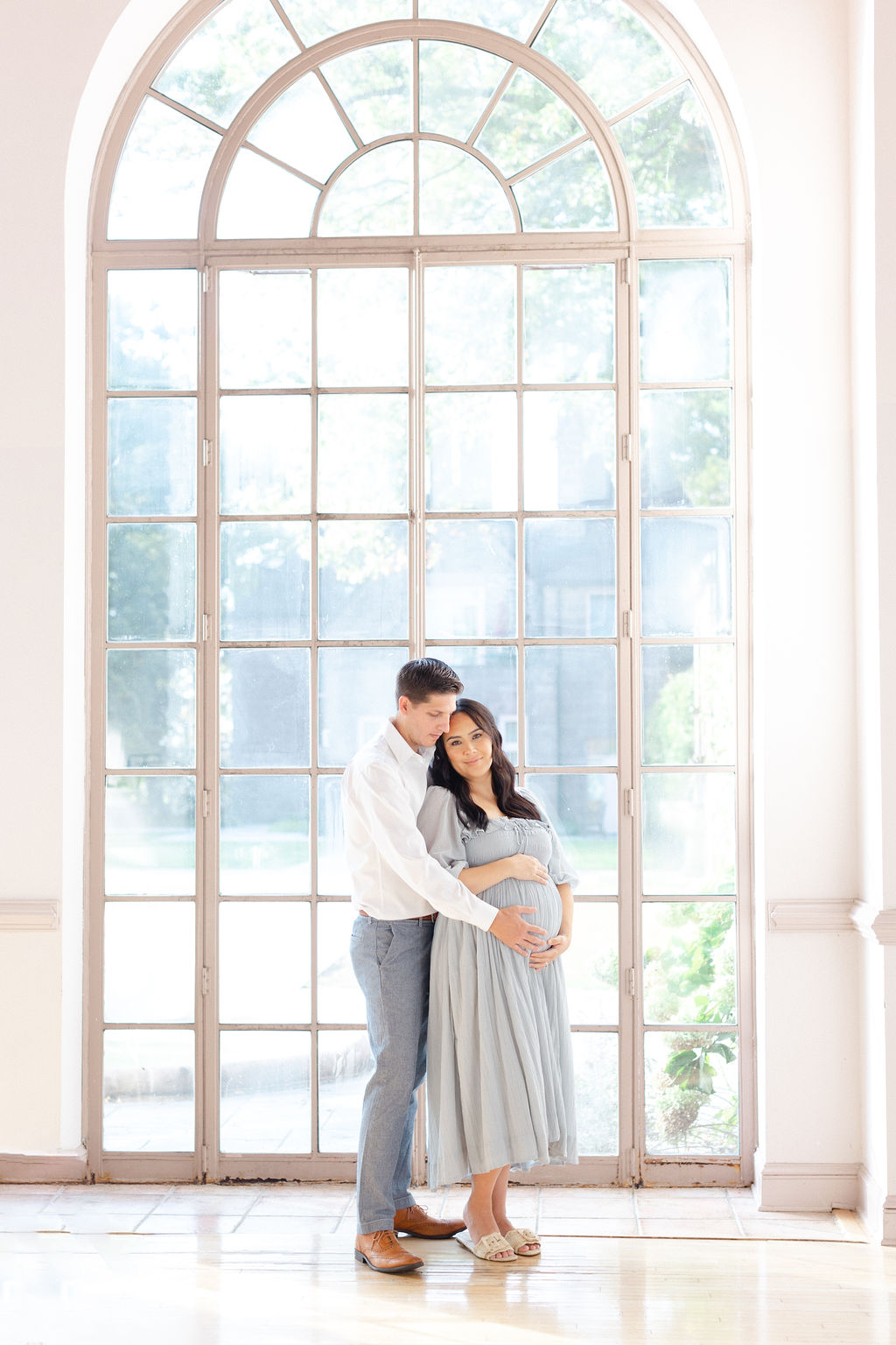 A mom to be stands in a large window leaning into the chest of her husband as they both hold the bump trimester fit body