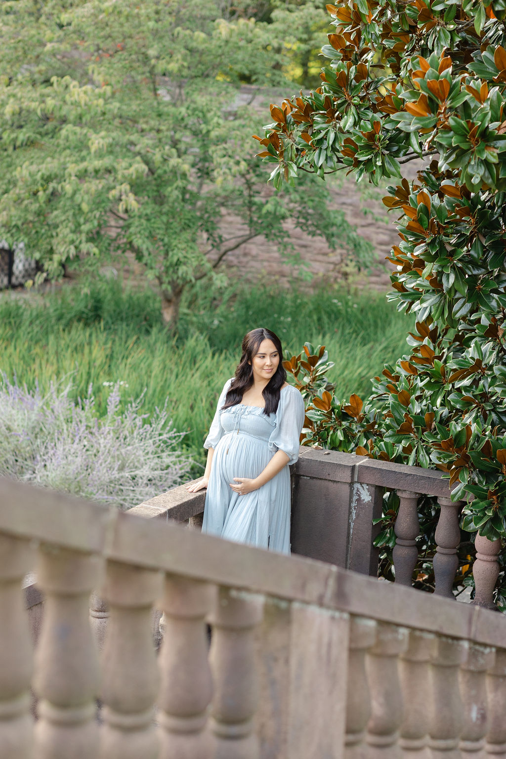 A mom to be in a blue maternity gown stands at the bottom of a large staircase looking over her shoulder and holding the bump by a magnolia tree after visiting trimester fit body
