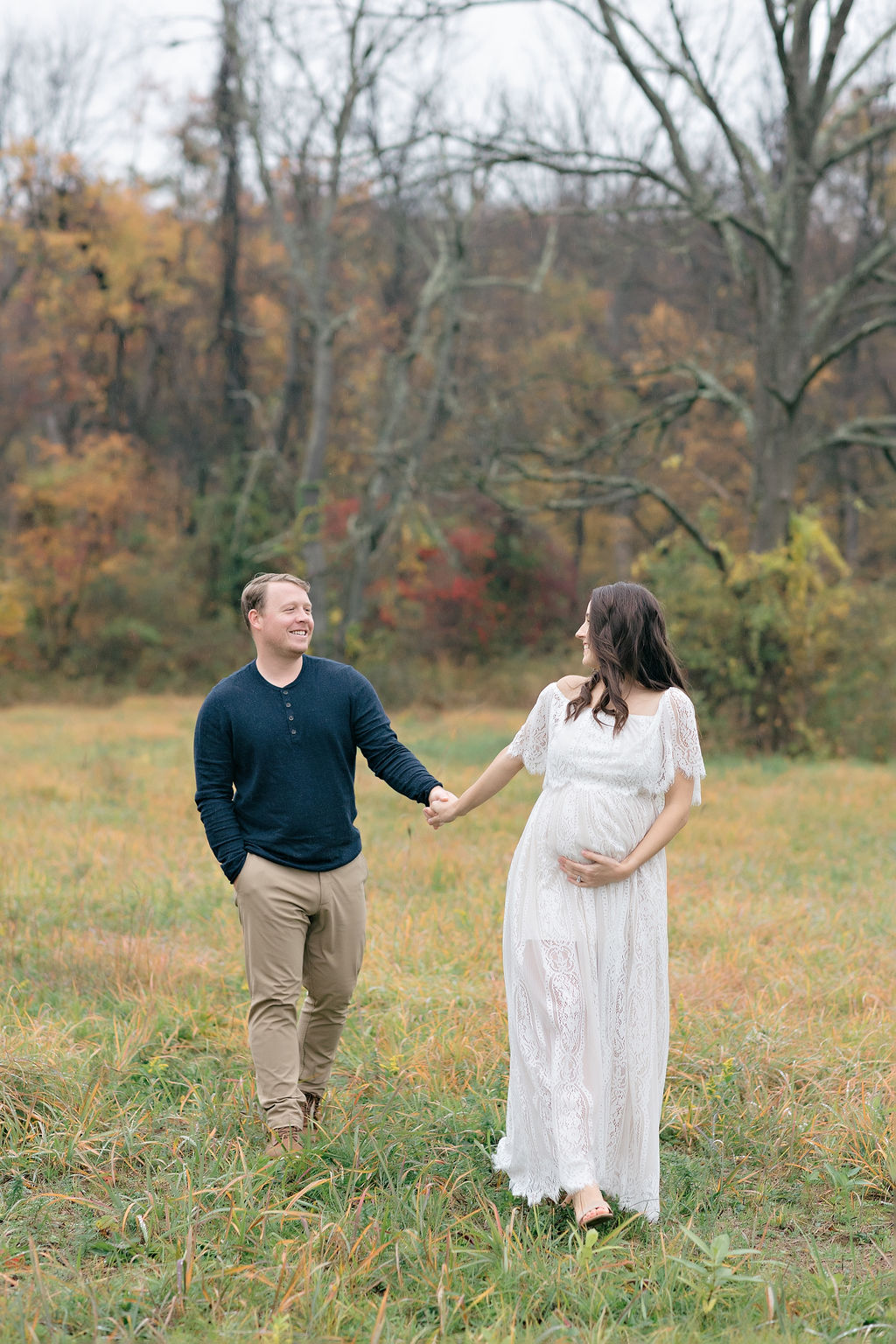 A mom to be in a white maternity gown leads her husband by the hand through a field of grass while holding the bump