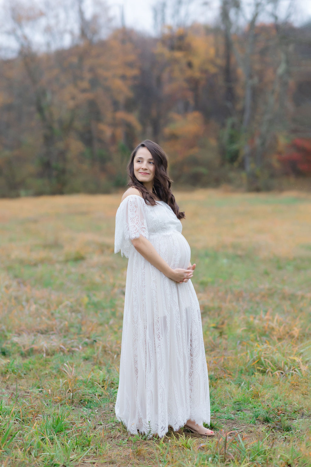 A mom to be in a white lace dress smiles over her shoulder while standing in a field holding her bump before getting a 4d ultrasound philadelphia