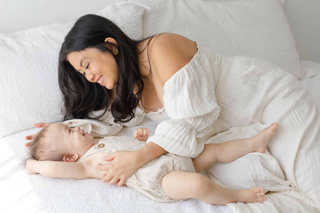 A mom in a white dress lays on a bed playing with her toddler son in matching overalls