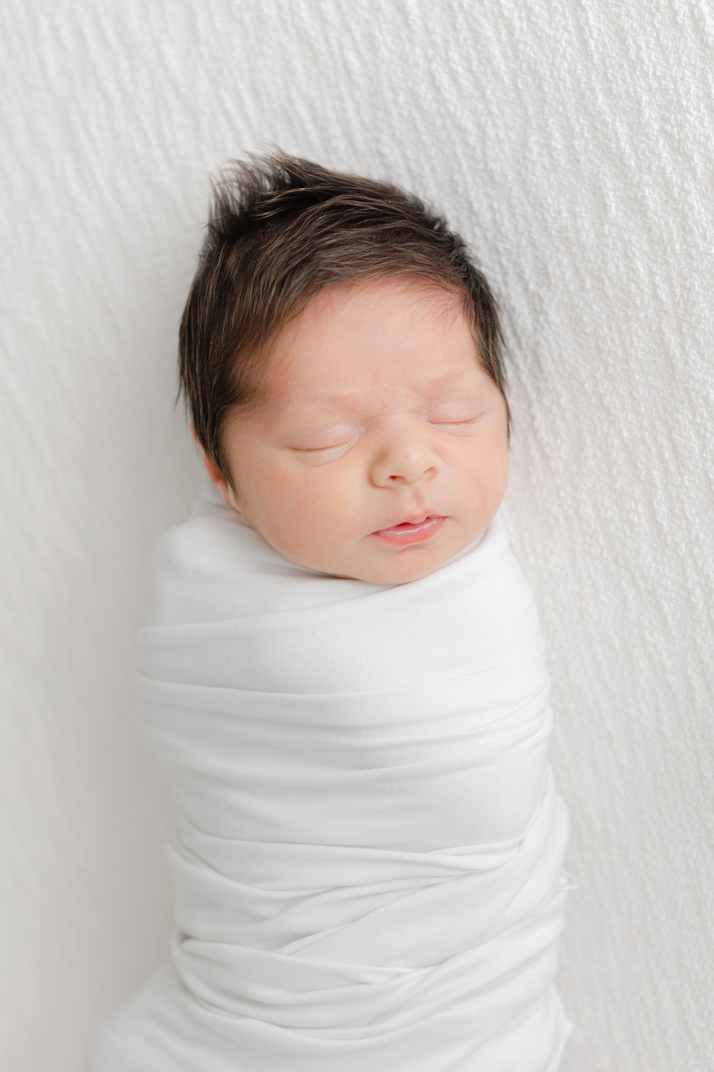 A newborn baby sleeps in a white swaddle on a white bed