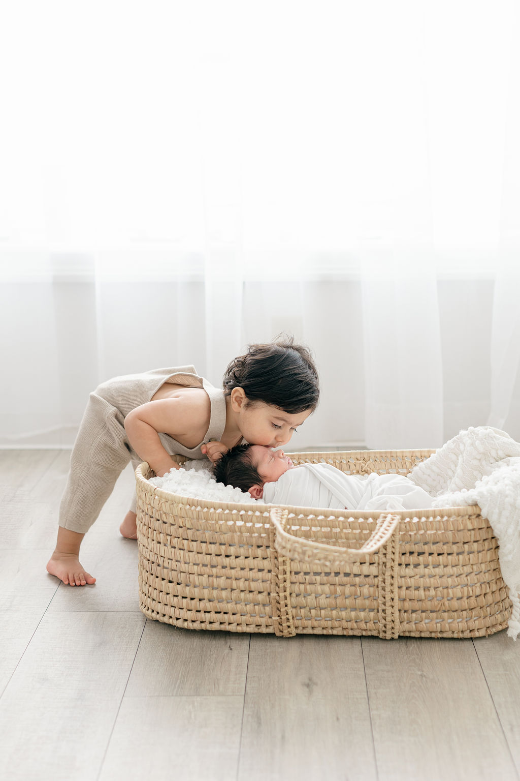 A toddler boy in tan overall kisses the head of his sleeping newborn sibling in a woven basket