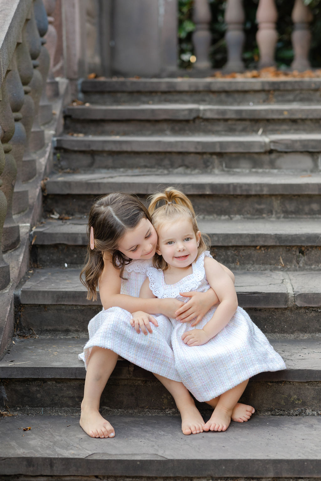 Two young toddler sisters sit on stone stairs hugging and smiling in matching white dresses before mom and dad have Philadelphia date nights