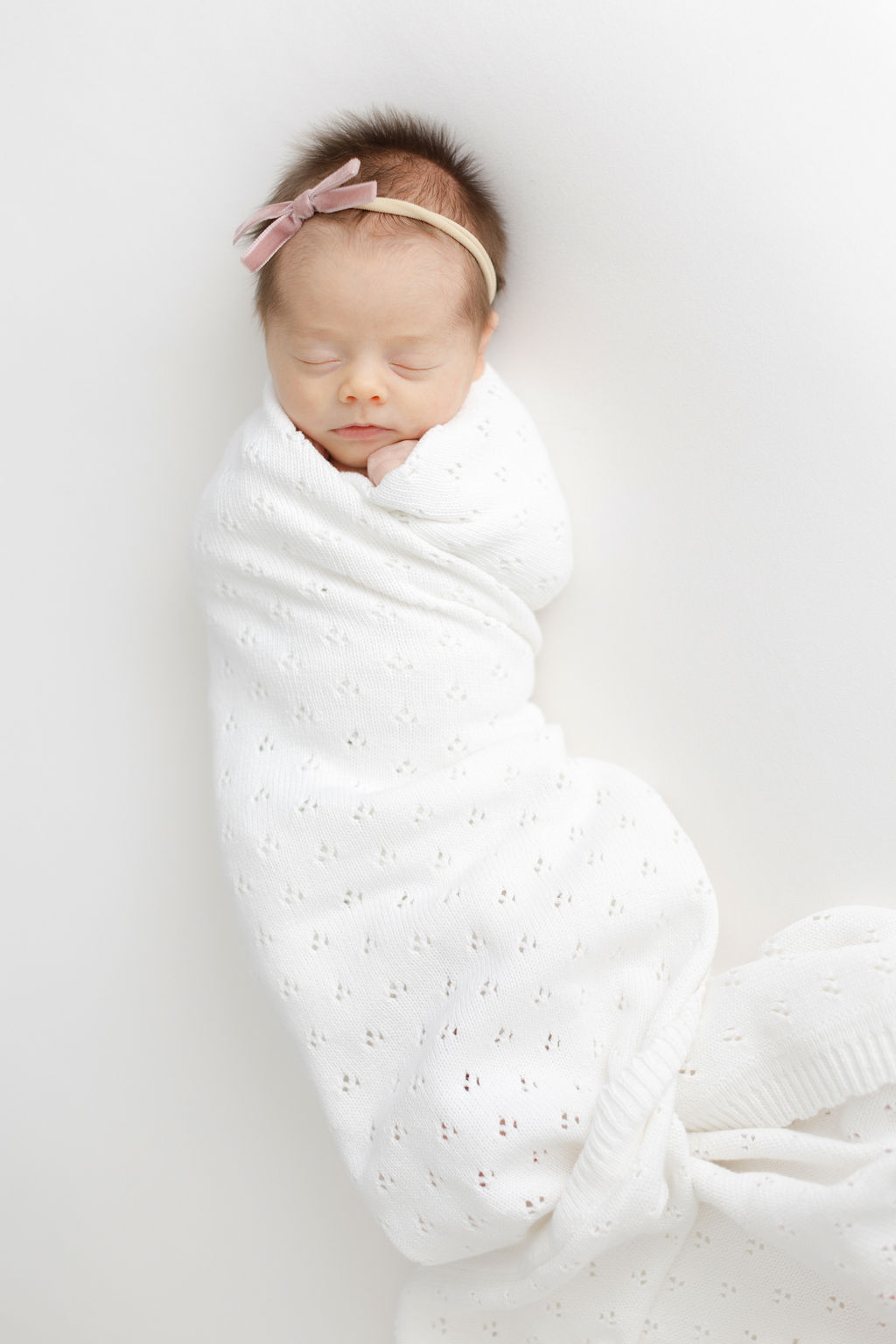 A newborn baby sleeps in a white blanket swaddle while wearing a pink bow headband