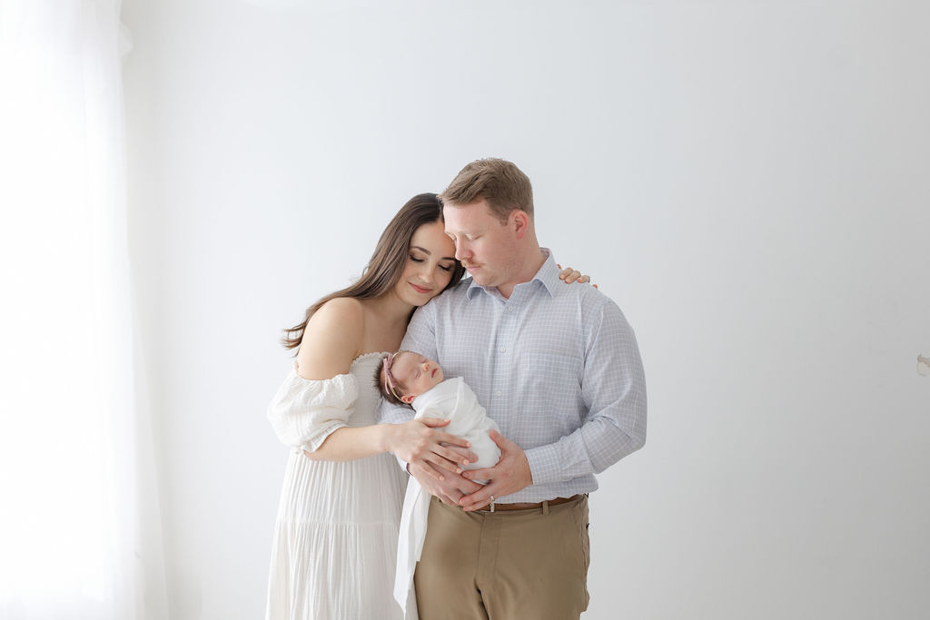 A mom and dad stand by a window in a studio smiling down to their newborn baby daughter in dad's hands after visiting a baby store princeton