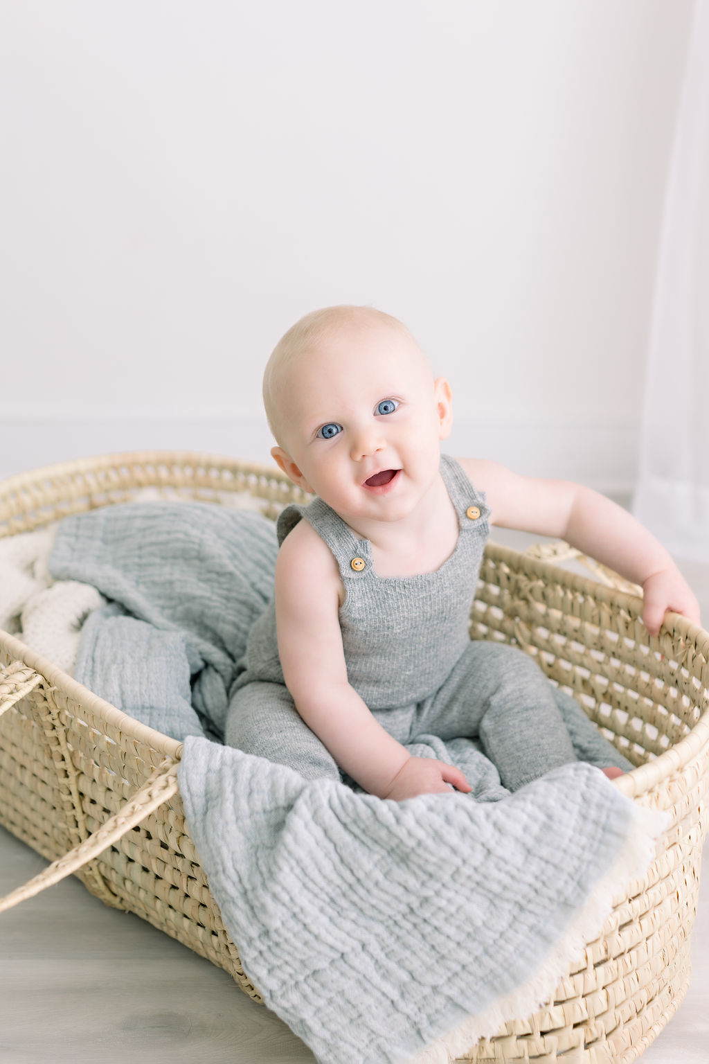 An infant boy in grey overalls plays while sitting in a woven basket in a studio after some baby swim classes Philadelphia