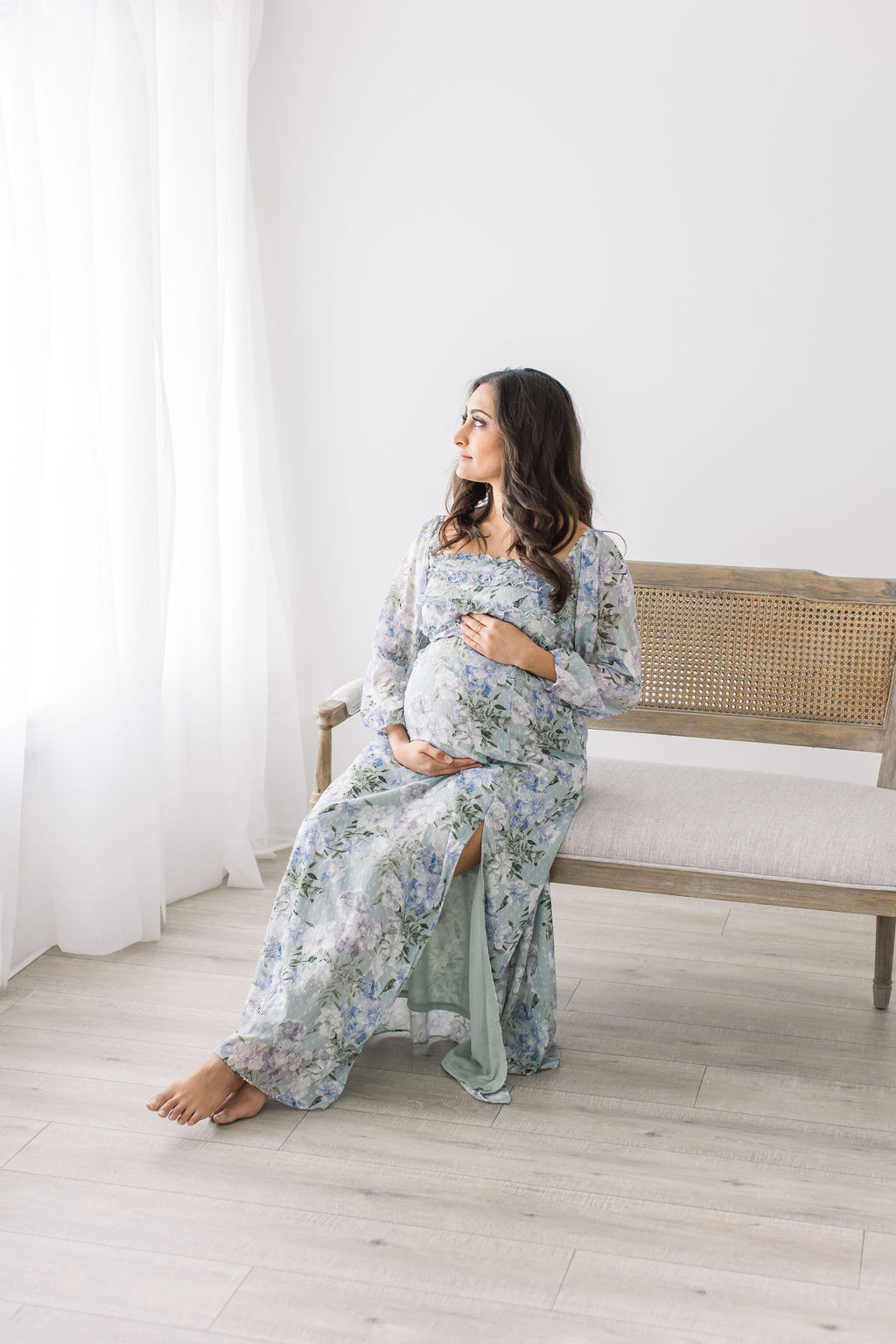 A mother to be sits on a bench in a studio holding her bump and looking out a window