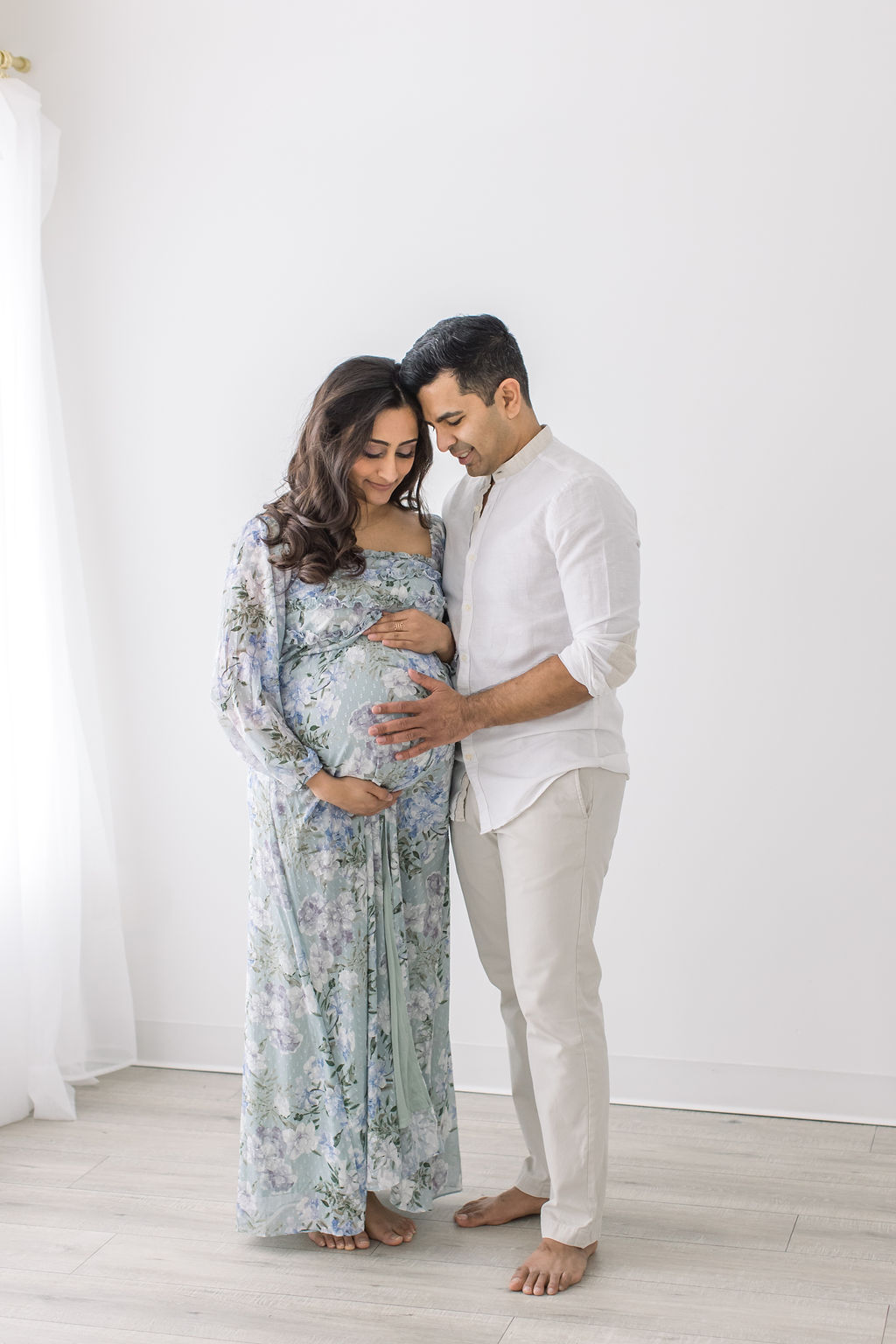 A pregnant mother in a floral dress stands in a studio smiling down to the bump with her husband after a prenatal massage princeton