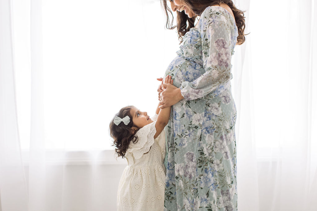 A pregnant mother stands in a studio wearing a floral dress and playing with her toddler daughter holding the bump after a prenatal massage princeton
