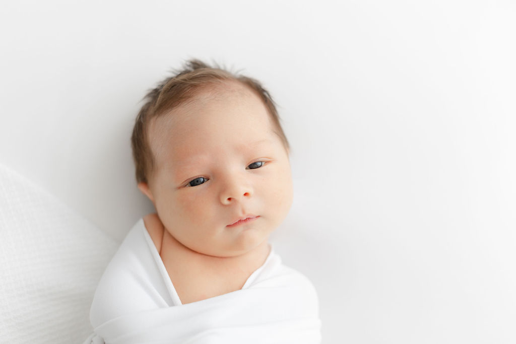 A newborn baby lays in a white swaddle with eyes open in a studio thanks to doulas princeton nj