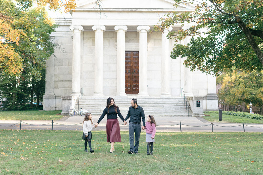 A mom and dad walk in a lawn in front of a classical building holding hands with their two young daughters