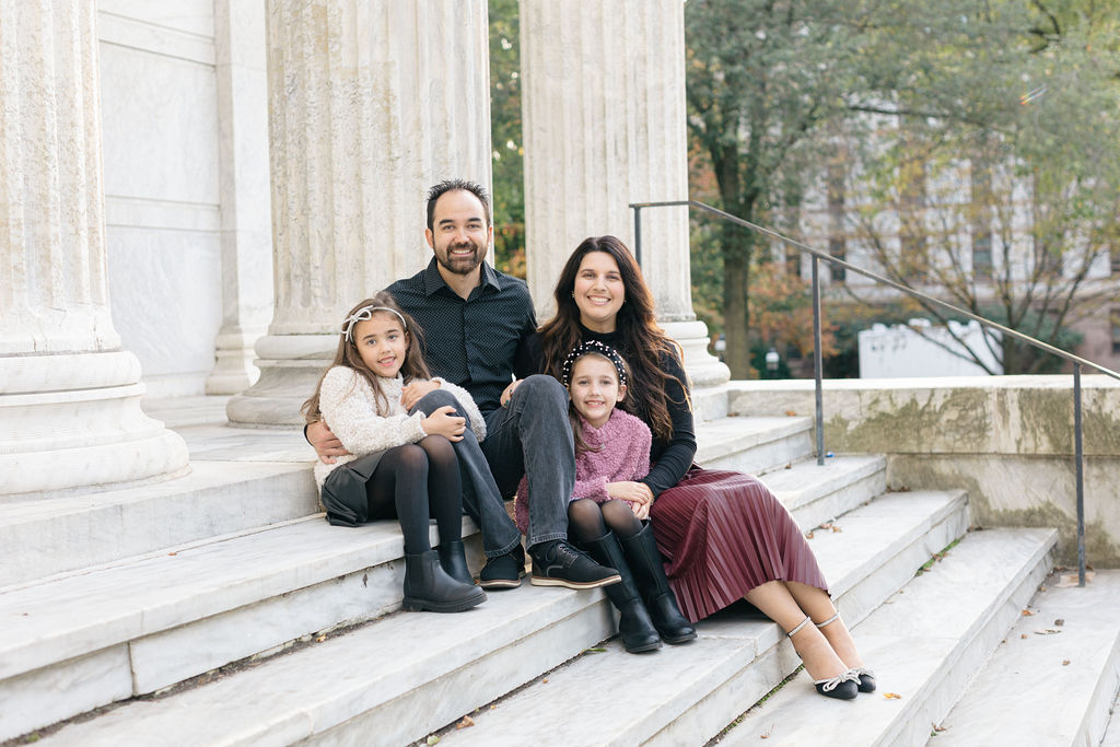 A mom and dad sit on some marble steps of a classical building with their two young daughters before visiting family friendly restaurants princeton