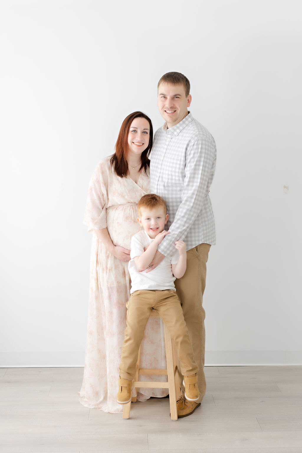 A mother and father stand in a studio with their young son sitting on a wooden stool in front of them