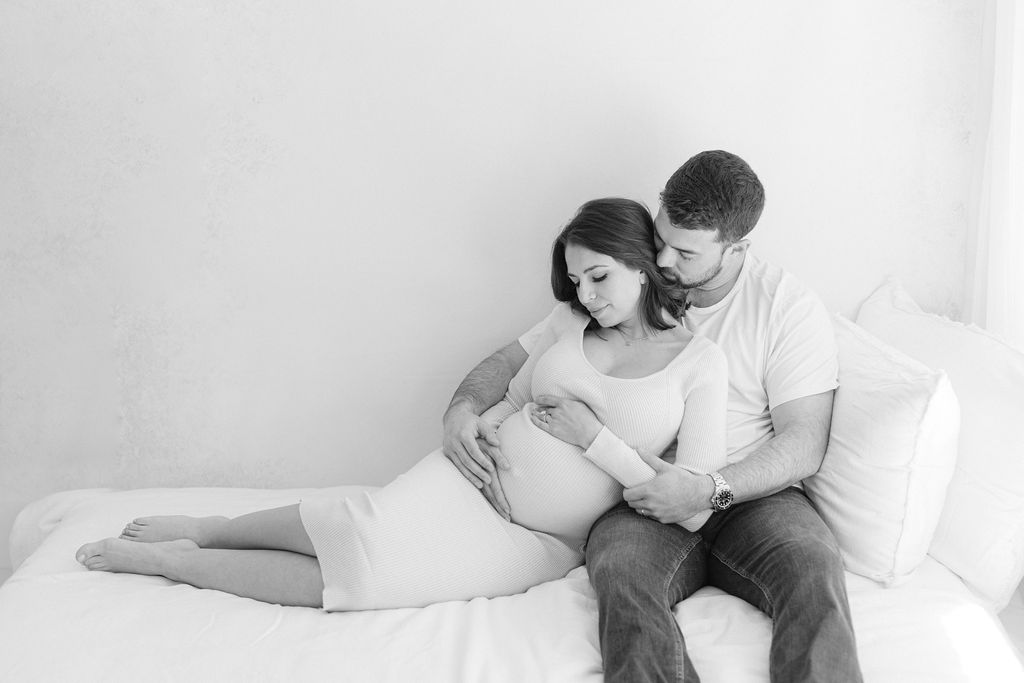 A mother to be leans into the chest of her husband while they sit on a bed in a studio