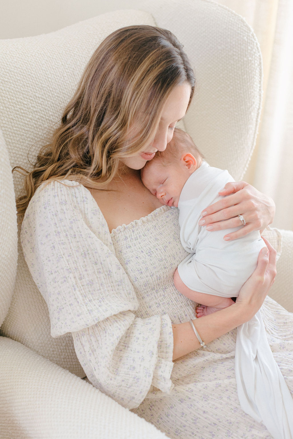 A mother sitting in a chair cradling her sleeping newborn against her cheek and chest as posed by a Philadelphia lifestyle newborn photographer