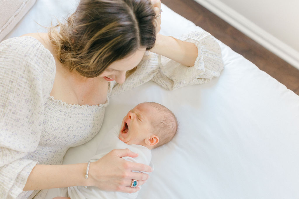 A mother in a floral print dress cuddles with her yawning newborn baby on a white bed during a session with a Philadelphia lifestyle newborn photographer