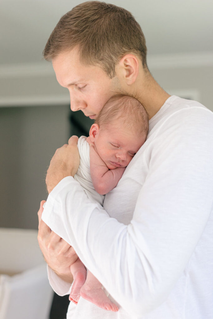 A new father in a white shirt cradles his sleeping newborn baby against his chest as guided by a Philadelphia lifestyle newborn photographer