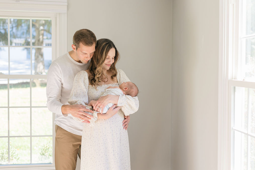 Happy new parents stand by some windows smiling down at their newborn baby in mom's arms