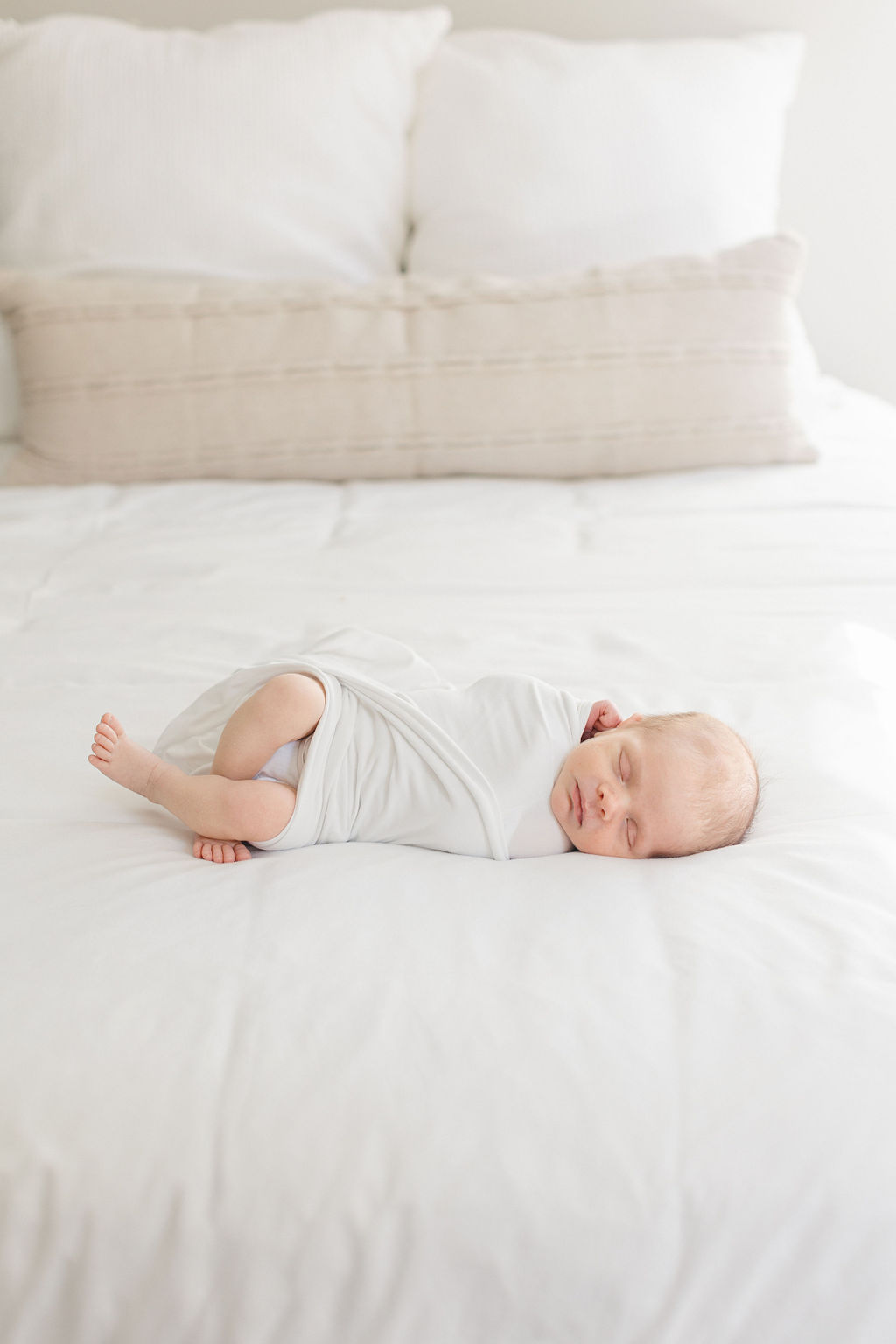 A newborn baby sleeps on its side in a white swaddle on a white bed