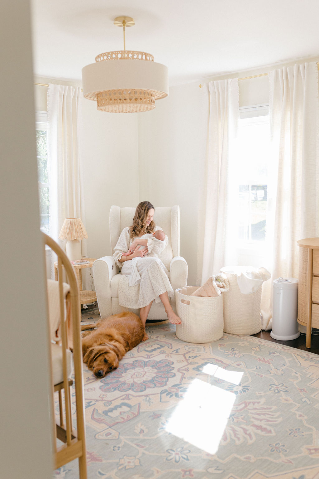 A mother sits in a nursery with her golden retriever laying at her feet and newborn baby in her arms for a Philadelphia lifestyle newborn photographer