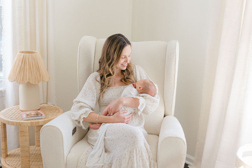 A happy mom sits in a nursing chair by a window with her sleeping newborn baby in her arms