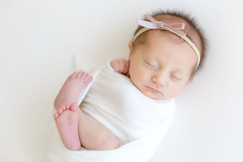 A newborn baby girl sleeps while wrapped in a right white swaddle with legs out and wearing a pink bow headband