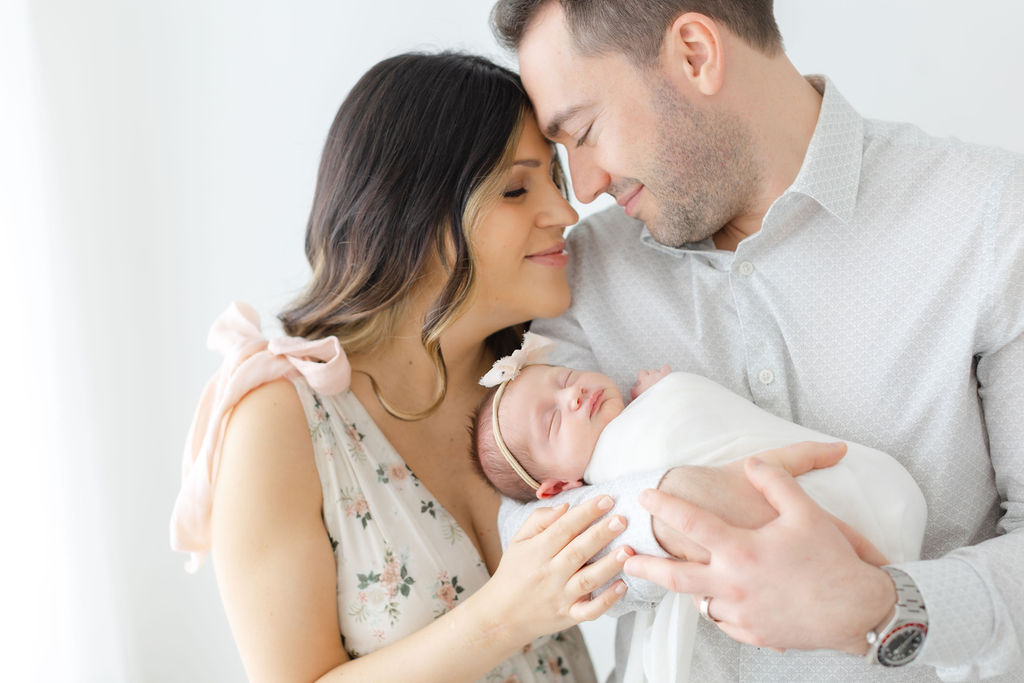 Happy new parents nuzzle foreheads while holding their sleeping newborn daughter in the studio of a bucks county newborn photographer
