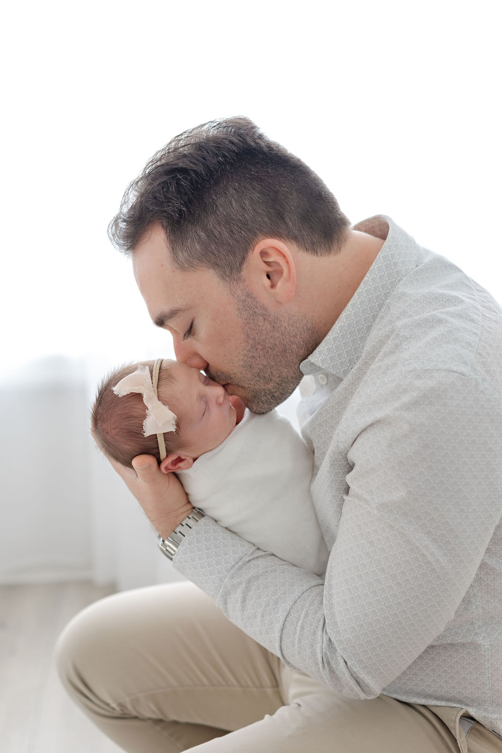 A new dad in a grey shirt sits under a window kissing his sleeping newborn baby daughter in the studio of a bucks county newborn photographer