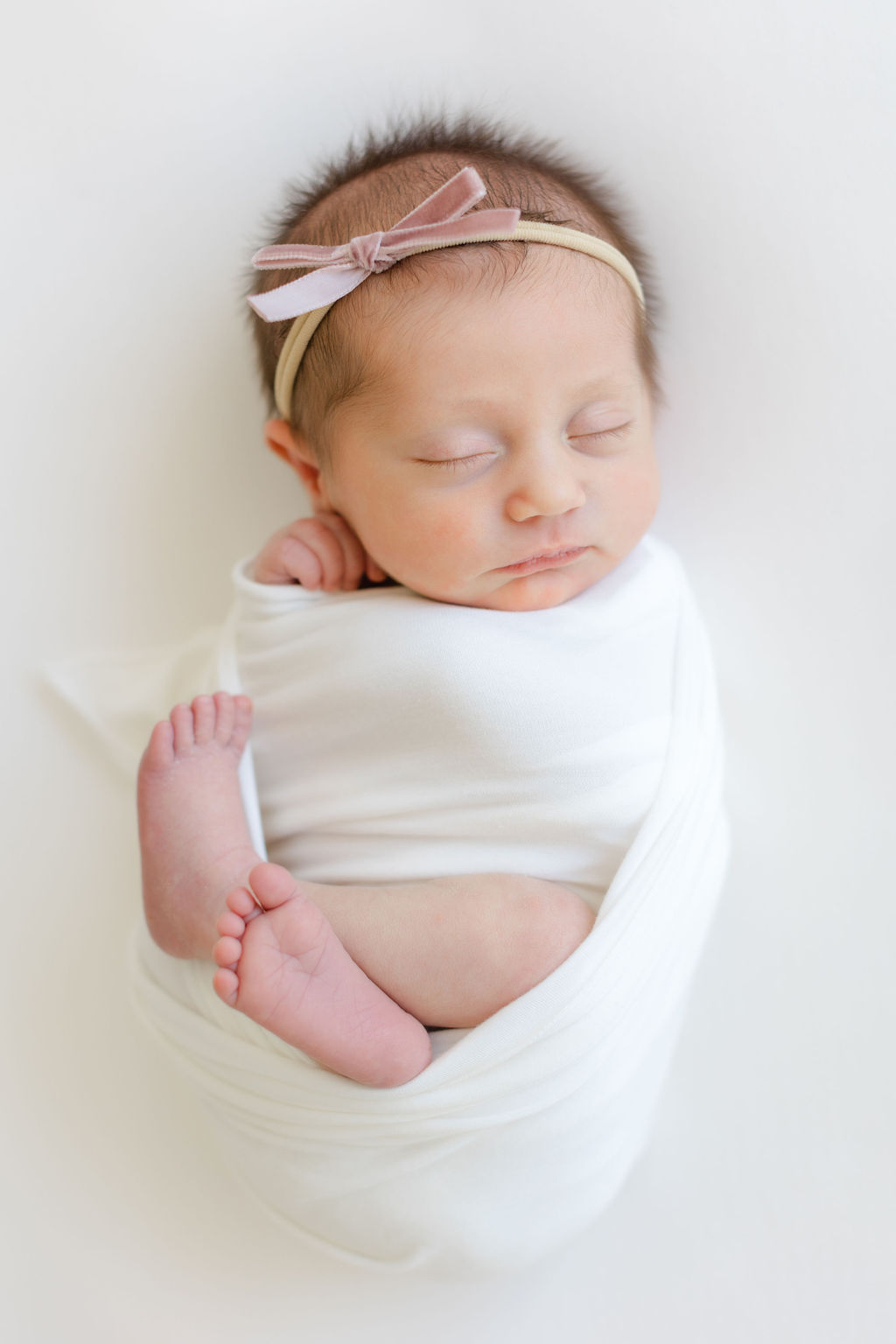 A newborn baby girl sleeps with a pink bow in a swaddle with legs sticking out