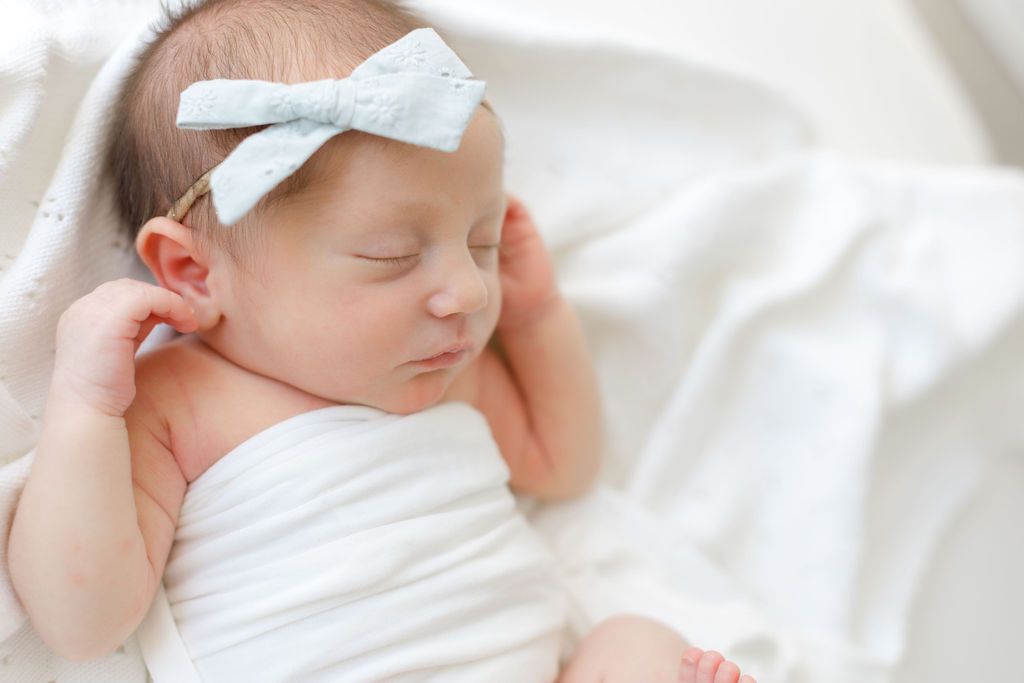 A newborn baby girl sleeps with a white headband bow in a white bed