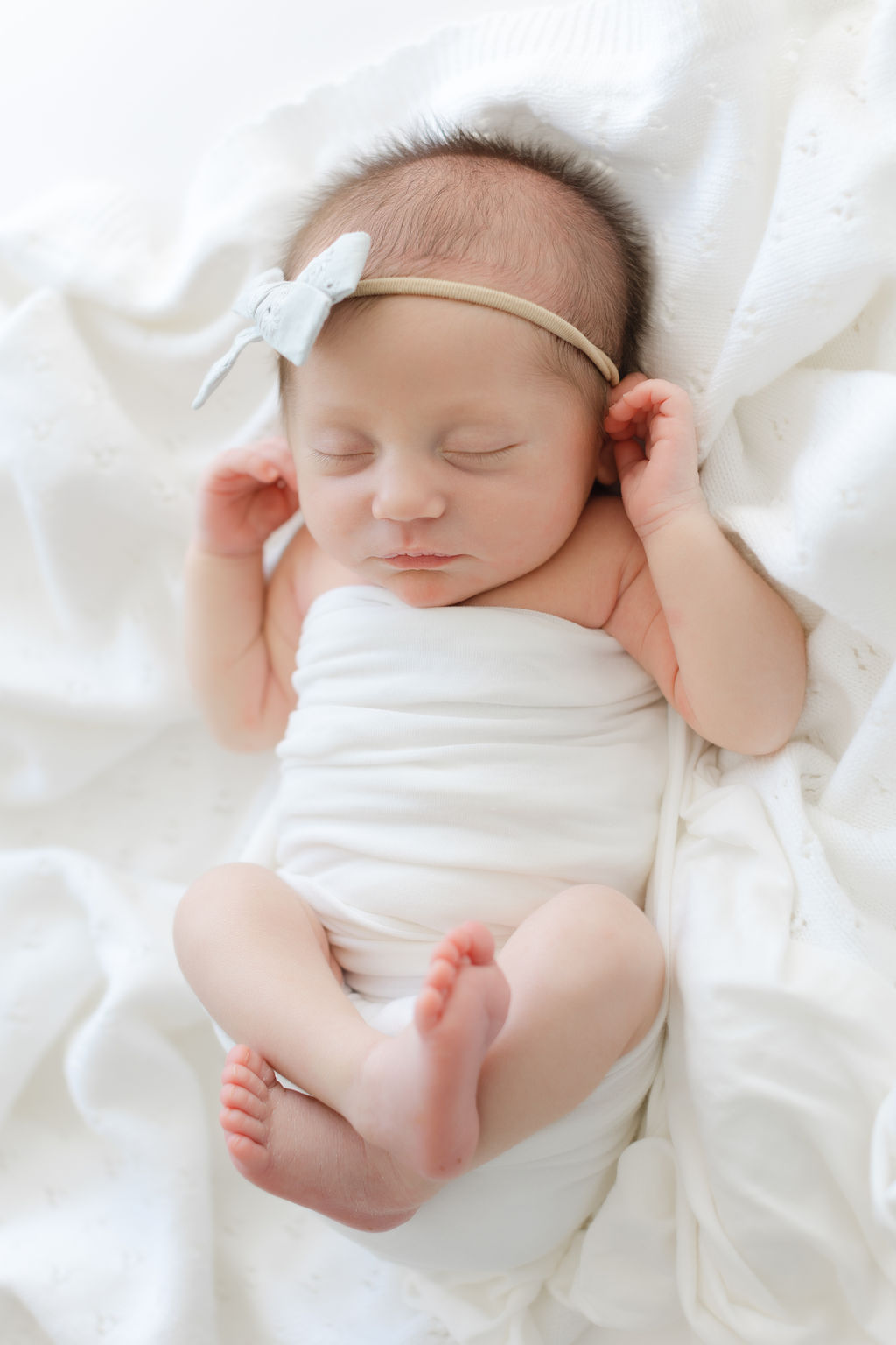 A newborn baby sleeps while wrapped with arms and legs loose in a white wrap on a white bed