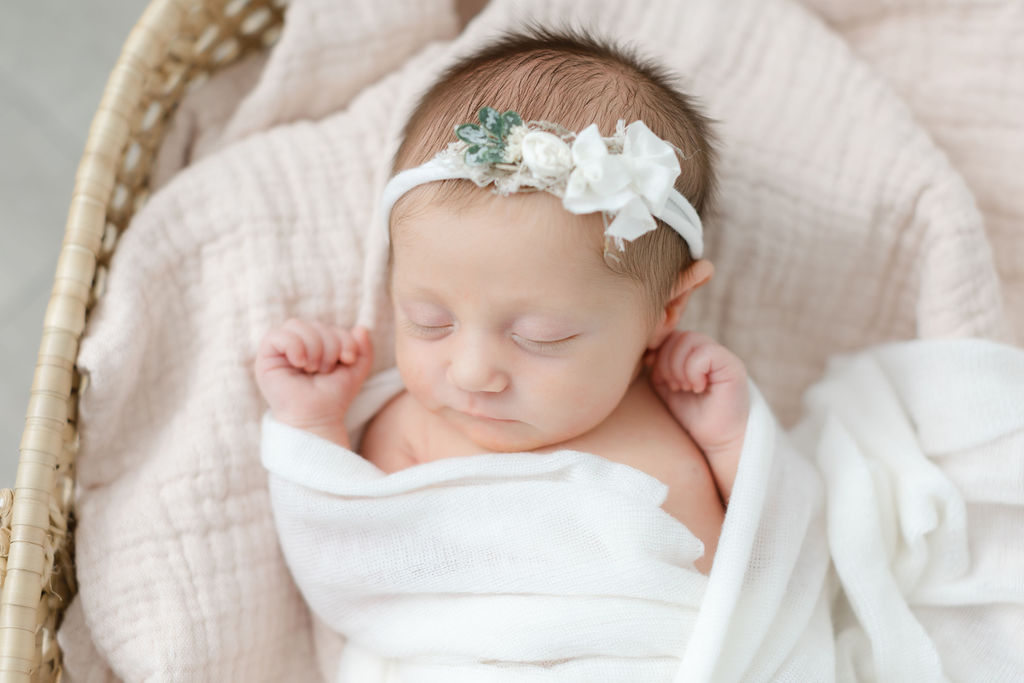 A newborn baby girl with a white bow headband and white swaddle sleeps in a woven basket in the studio of a bucks county newborn photographer