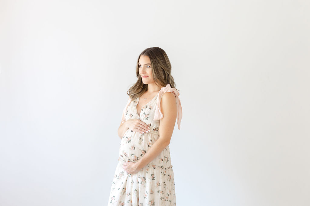 A happy mom to be stands in a Doylestown Maternity photos studio wearing a white floral maternity gown smiling