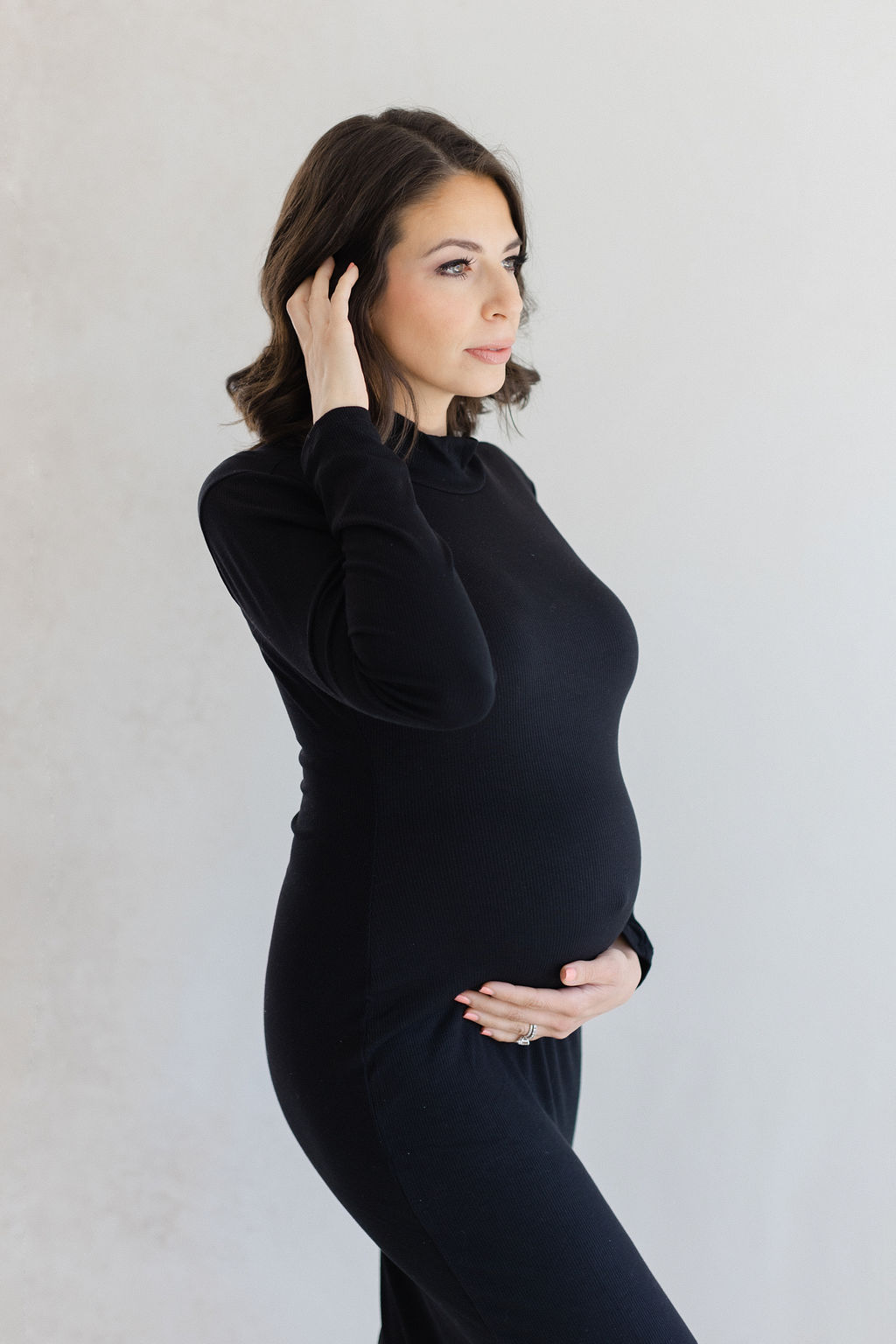 A pregnant woman in a black maternity gown stands in a Doylestown Maternity studio holding her bump