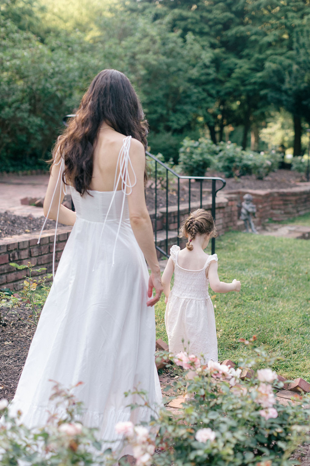 A mother and daughter in white dresses wander through a flower garden