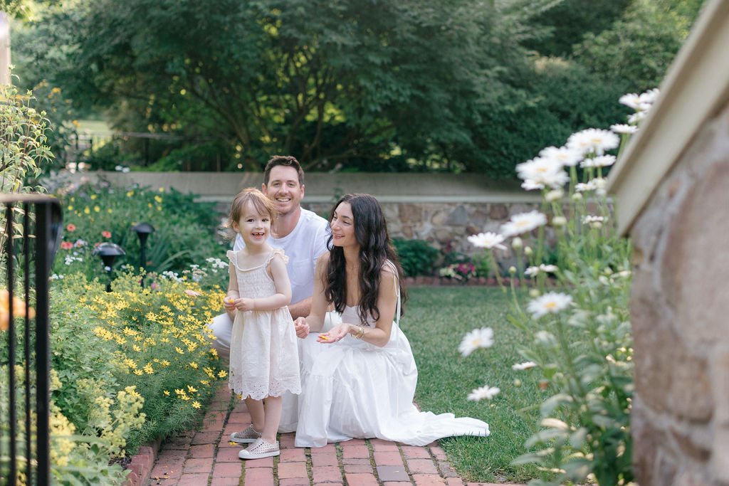 A mom and dad in white explore a flower garden with their toddler daughter before some ice cream bucks county